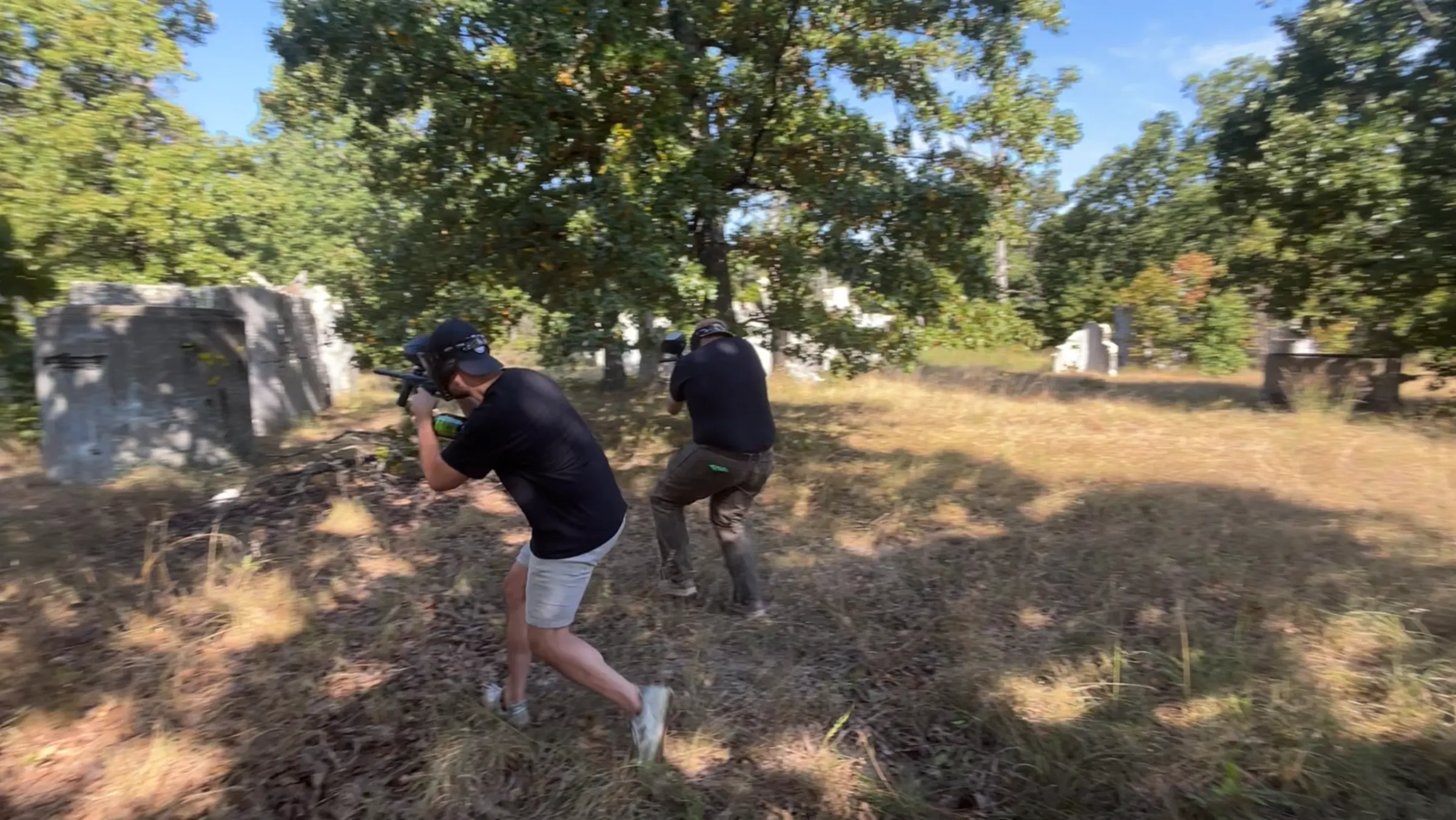 Two paintball players advancing through a wooded field past concrete bunkers